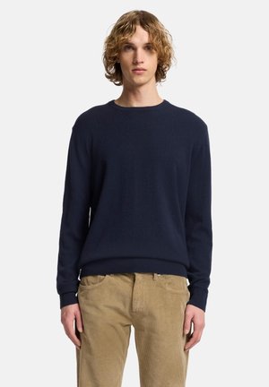 Giovane con capelli ricci fino alle spalle, che indossa un maglione blu navy a maniche lunghe e pantaloni beige in velluto a coste, in piedi rivolto verso l'obiettivo.