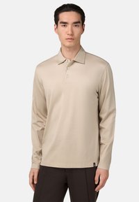 Beige polo majica z dolgimi rokavi in klasičnim ovratnikom, z zapenjanjem na tri gumbe in mehkim materialom; v kombinaciji s temnimi hlačami; minimalne podrobnosti.