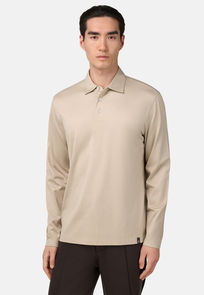 Beige polo majica z dolgimi rokavi in klasičnim ovratnikom, z zapenjanjem na tri gumbe in mehkim materialom; v kombinaciji s temnimi hlačami; minimalne podrobnosti.