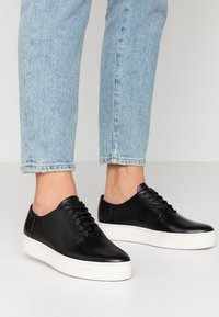 Zapatillas deportivas de cuero negro con textura suave, con una suela de goma blanca y seis ojales con cordones. Diseño simple y de perfil bajo.