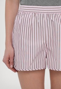 Femme portant un short à rayures verticales roses, blanches et marron avec une taille élastique et un haut gris, main posée sur le côté.