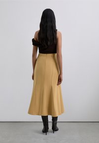 Femme aux longs cheveux noirs portant un haut noir sans épaule, une jupe midi beige et des bottes à talons noires, se tenant face à un fond clair uni.