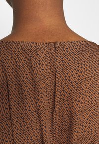 Tissu marron à motifs avec des pois navy, doté d'une texture douce et d'un dos avec une encolure agrémentée d'une petite fermeture à bouton au cou.