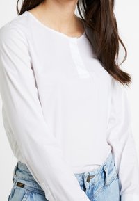 Chemise blanche à manches longues avec un col rond et une petite patte de boutonnage à l'avant. Tissu lisse, silhouette ajustée, portée avec un jean taille haute bleu clair.