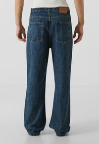 Bredde ben blå denimjeans med sømløs bakside, to lommer og en brun lærlogo ved livet.