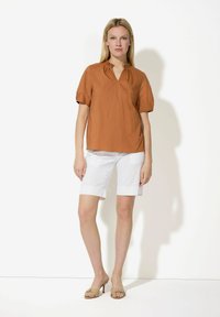 Blouse courte marron à manches courtes avec décolleté en V et détails plissés, associée à des shorts blancs à revers et des sandales à talons de couleur neutre.