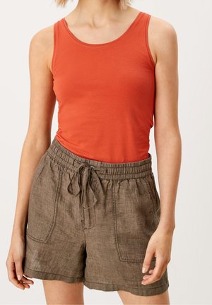 Mujer con top sin mangas naranja y pantalones cortos marrones sueltos con cordón, de pie frente a un fondo liso y claro.