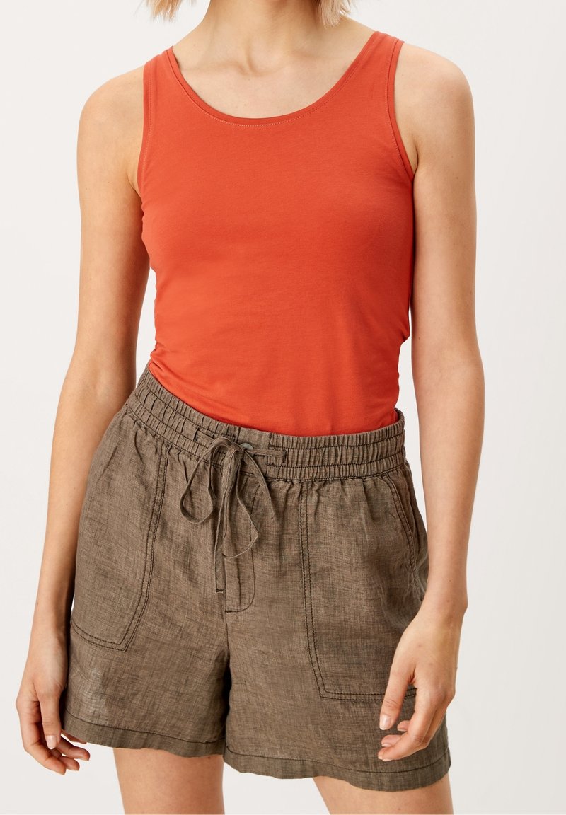 Femme portant un haut orange sans manches et un short marron ample à cordon, debout devant un fond clair uni.