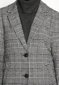 Blazer a cuadros en gris y negro con dos botones negros, usado sobre un suéter de cuello alto en gris oscuro con textura de canalé.