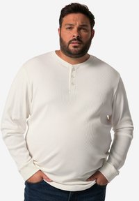 Weißes Langarm-Henley-Shirt aus strukturiertem Stoff, mit Knöpfen vorn und gerippten Bündchen. Getragen mit dunkelblauen Jeans. Einfaches Design und neutrale Farbe.
