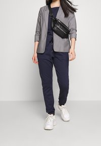 Grå blazer över en marinblå jumpsuit, matchad med en svart crossbody-väska och vita sneakers. Slät tyg, skräddarsydd passform, elastiska manschetter.