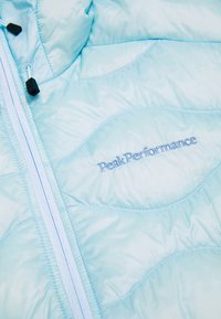 Lys blå isoleret jakke lavet af skinnende materiale, med en fuld lynlås, dekorativ syning i et bølgemønster, og "Peak Performance" logo.