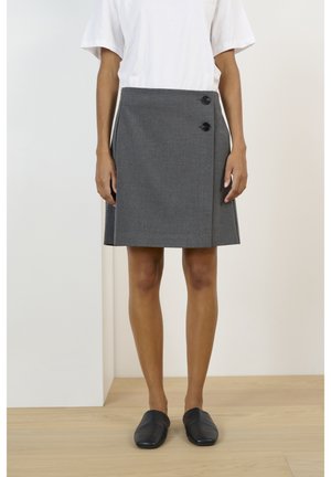 Person iført en grå nederdel til knæet med to sorte knapper, kombineret med en hvid top og sorte slip-on sko, stående på et trægulv.