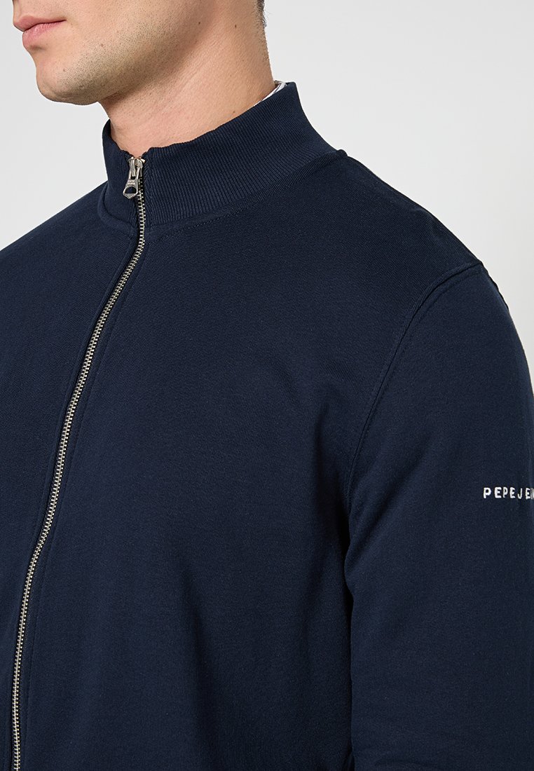 Mann trägt einen marineblauen Reißverschluss-Pullover mit geripptem Kragen und dem aufgenähten "Pepe Jeans"-Logo auf dem linken Ärmel.