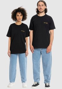 Zwarte katoenen t-shirts met gouden logo, losse pasvorm. Gecombineerd met lichtblauwe spijkerbroeken. Eén persoon draagt witte sneakers, de andere zwarte.
