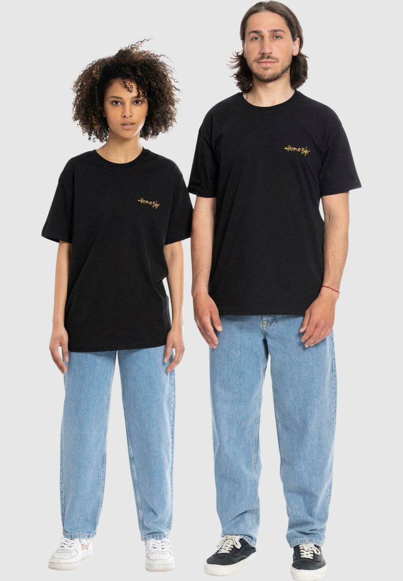 Zwarte katoenen t-shirts met gouden logo, losse pasvorm. Gecombineerd met lichtblauwe spijkerbroeken. Eén persoon draagt witte sneakers, de andere zwarte.
