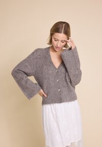 Grå, fluffy cardigan med lange ermer og v-hals, med fem knappelukninger og en myk tekstur. Bærer over et hvitt skjørt.