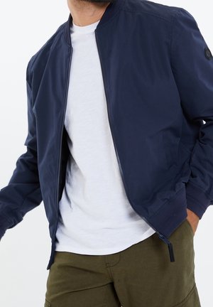 Homme portant une veste aviateur bleu marine sur un t-shirt blanc et un pantalon vert olive, une main dans la poche, torse visible.