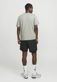 Grå kortærmet skjorte, sorte shorts, hvide sokker med tekst. Model vender bort, iført sølvfarvede sneakers, der viser et simpelt atletisk design.