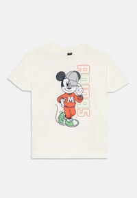 Camiseta crema con un ratón de dibujos animados vestido con un chándal naranja, zapatillas verdes y una gorra, acompañado de un colorido texto "ADIDAS".