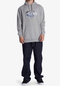 Persoon die een grijze hoodie met een grafisch ontwerp draagt, donkerblauwe jeans en zwarte sneakers, staand tegen een witte achtergrond.