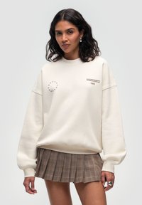 Felpa off-white con spalle scese, caratterizzata da dettagli ricamati e un motivo circolare di stelle. Abbinata a una mini gonna a quadri marrone.