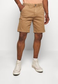 Shorts en coton kaki avec poches avant, fermeture à bouton et ourlets retroussés, accompagnés de baskets montantes blanches.