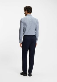 Lichtblauw geruit overhemd met lange mouwen, gecombineerd met marineblauwe broek en zwarte schoenen, gezien vanaf de achterzijde. Knopen op de manchetten zichtbaar.