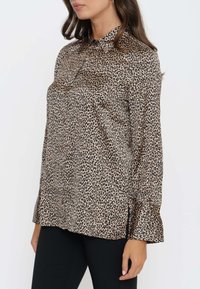 Blusa in stampa animale in misto seta beige e nera. Presenta un collo a camicia, chiusura con bottoni, maniche lunghe e una vestibilità rilassata con spacco laterale.