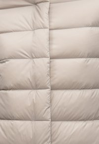 Svetlo béžová prešívaná bunda s prešitými časťami, hladkou textúrou a jemným leskom. Má zvislé prešívanie a rovnomerné rozloženie výplne.