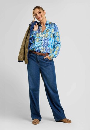 Donna con camicia blu a motivi, jeans a gamba larga, che tiene una giacca trapuntata marrone sulla spalla, in piedi con una mano in tasca.