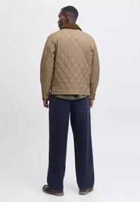 Hombre de espaldas vistiendo una chaqueta acolchada color crema con un cuello marrón, pantalones amplios azul marino y zapatos marrones, de pie contra un fondo blanco liso.