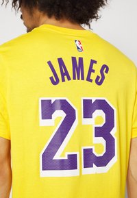 Gul kortermet T-skjorte i bomull med stort lilla tall "23" og navnet "JAMES" på baksiden, med NBA-logo etikett ved halsen.