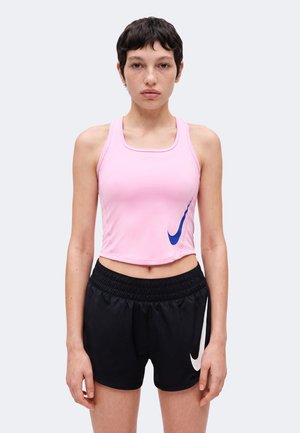 Jeune femme aux cheveux courts et foncés portant un crop top rose sans manches et un short de sport noir avec des logos Nike, debout devant un fond uni.