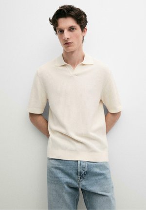 Junger Mann mit kurzen lockigen Haaren, der ein cremefarbenes, geripptes Kurzarm-Poloshirt und hellblaue Jeans trägt und an einer weißen Wand steht.