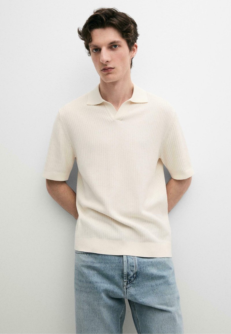 Jeune homme aux cheveux courts et bouclés portant un polo côtelé crème à manches courtes et un jean bleu clair, debout contre un mur blanc.