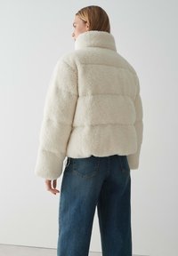 Giacca a vento fluffy color crema con colletto alto, caratterizzata da sezioni trapuntate orizzontali; abbinata a jeans a gamba larga blu scuro.