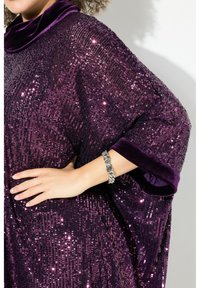 Poncho viola con paillettes e colletto in velluto. Presenta una vestibilità ampia e una texture lucida, accompagnato da un braccialetto argentato su una mano.