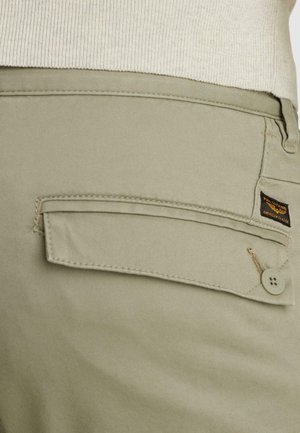 Pantalones beige con solapa de bolsillo trasero abotonada y una pequeña etiqueta de marca "American Classic" en negro y amarillo.