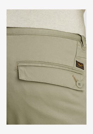 Pantaloni beige con battente della tasca posteriore chiuso da bottone e piccola etichetta del marchio "American Classic" in nero e giallo.