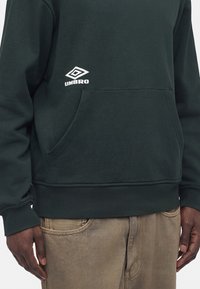 Άτομο που φοράει σκούρο πράσινο φούτερ Umbro με μπροστινή τσέπη και μπεζ παντελόνι, ορατό από το μέσο του στήθους μέχρι τα χέρια.