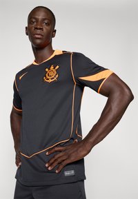 Svart atletisk drakt med oransje aksenter og kanting, med en logo på brystet. Laget av glatt, pustende stoff.