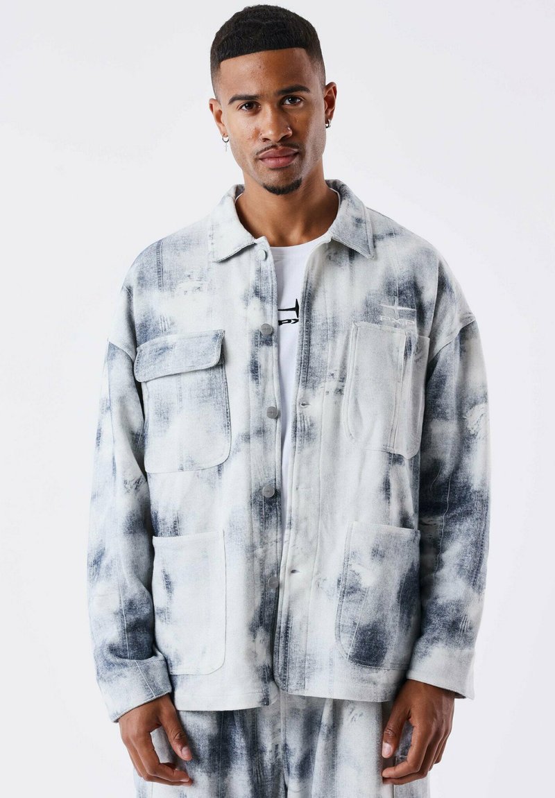 Veste à boutons en motif tie-dye blanc et gris, avec deux poches avant, une coupe décontractée et une texture douce. Portée par-dessus une chemise blanche.
