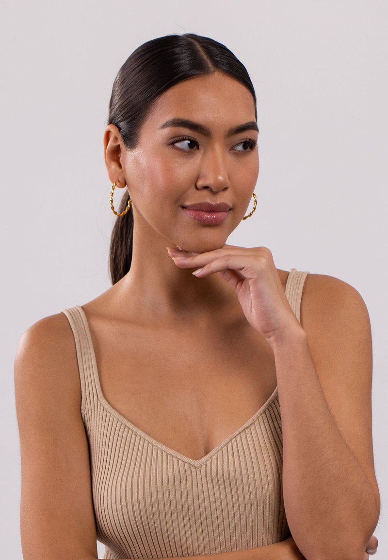 Beige gerippter Tanktop mit dünnen Trägern und V-Ausschnitt; das Model trägt goldene Creolen und hat glattes, dunkles Haar.