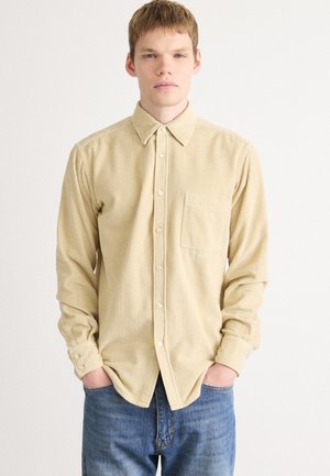 Light beige corduroy shirt with a button-down collar, long sleeves, front pocket, and textured surface; paired with blue jeans.