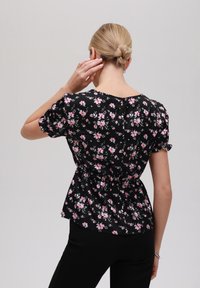 Svart blommig blus med rosa blommor, korta puffärmar, peplummidja och rund halsringning. Slätt tyg med en subtil glans.