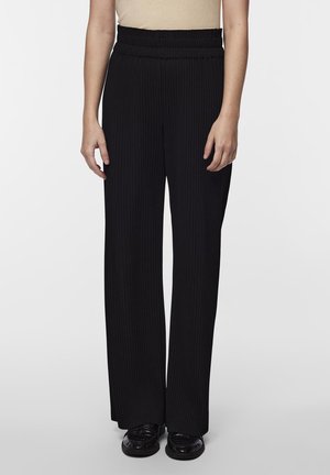 Pantalons larges noirs avec une texture côtelée, dotés d'une taille haute élastique et d'une coupe droite. Portés avec des chaussures plates noires.