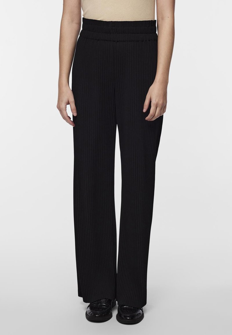 Pantalons larges noirs avec une texture côtelée, dotés d'une taille haute élastique et d'une coupe droite. Portés avec des chaussures plates noires.