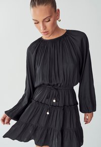 Crna haljina dugih rukava s naboranim izrezom, strukom na vezivanje i s tri slojeva ruffles na rubu; mekani, lepršavi materijal.