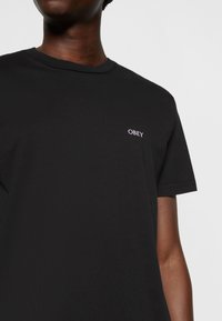 Černé bavlněné tričko s kulatým výstřihem, které má malé bílé logo "OBEY" na levém horním prsním místě, krátké rukávy a hladkou texturu.